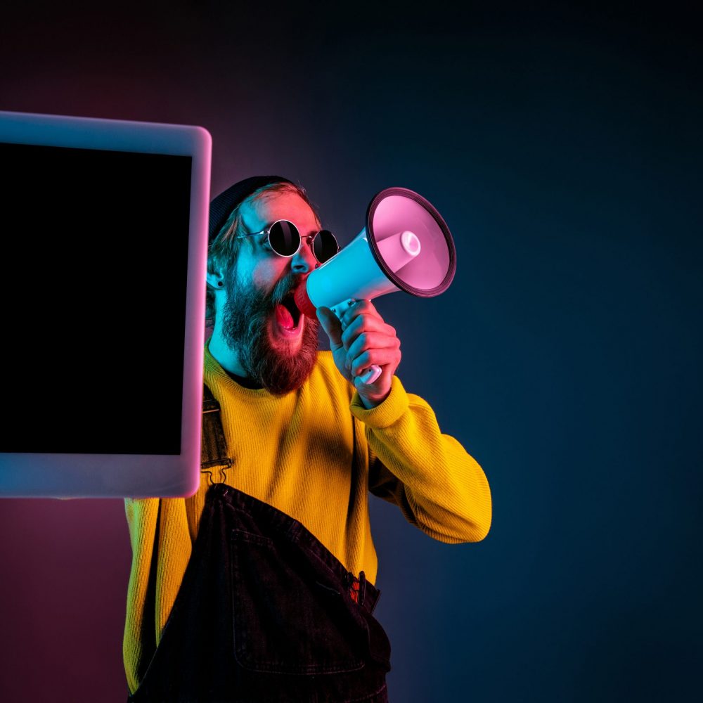 Showing tablet's blank screen. Caucasian man's portrait on gradient studio background in neon light. Beautiful male model with hipster style. Concept of human emotions, facial expression, sales, ad.